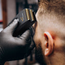 A close-up of a black beard trimmer with gold-colored blades being used by a person wearing a black glove. The brand name 'AIRDUTY' is clearly visible on the trimmer.